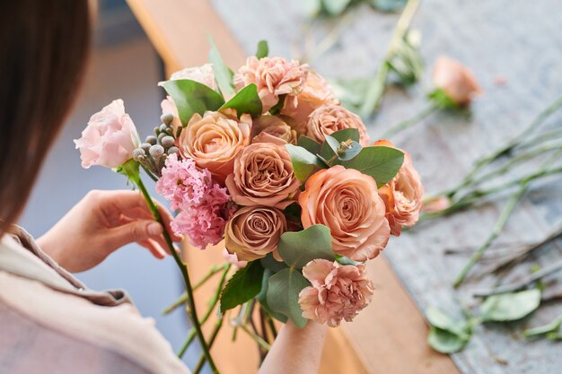 bunch of fresh roses and carnations of pastel colors held by young florist making bouquets by workplace