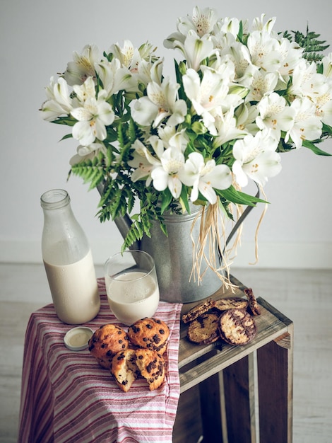 Bunch of fresh white flowers in metal vase placed near delicious muffins and milk on crate with striped cloth in studio