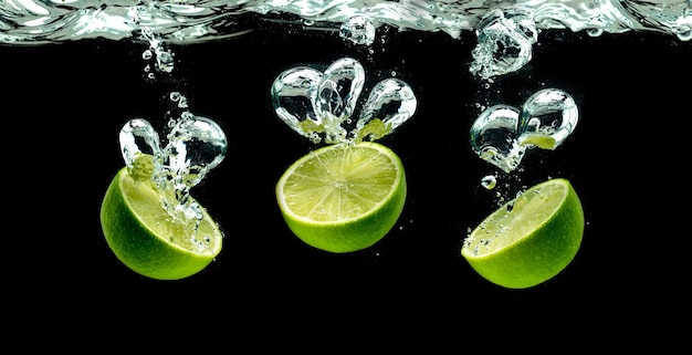 Bunch of lime fruits halves splashing with bubbles into water isolated against black background