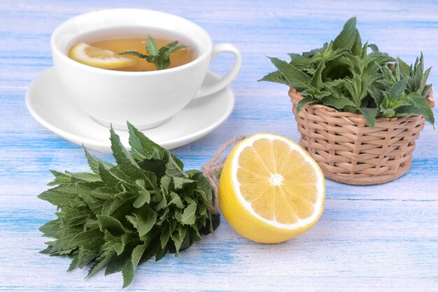 A bunch of mint with a lemon, a basket and a white cup of tea on a blue wooden background.