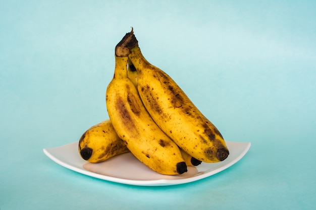 Букет из спелых бананов на белой тарелке на синем фоне. Крупным планом фото фонда.