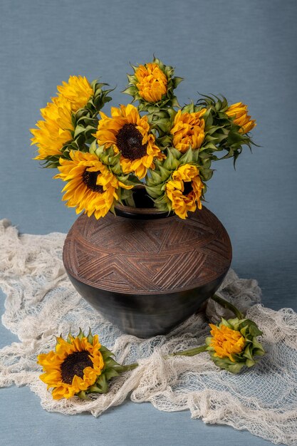 A bunch of yellow sunflowers in a clay pot on a blue background and a brown table. Fabric drapery.