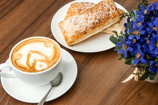 Buns in a plate and a cup of coffee with the spoon on a brown table with a flower