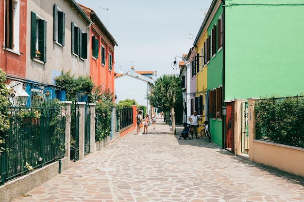 Burano, Venice, Italy - July 2, 2018: Panoramic view of brightly coloured homes of Burano is an island in the Venetian Lagoon. People walk and rest on streets. Summer sunny day and blue sky