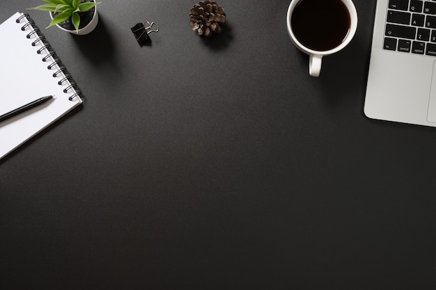 Foto bureaukantoor bovenaanzicht met toetsenbord, pen, notebook, koffie en plant op zwarte achtergrond.