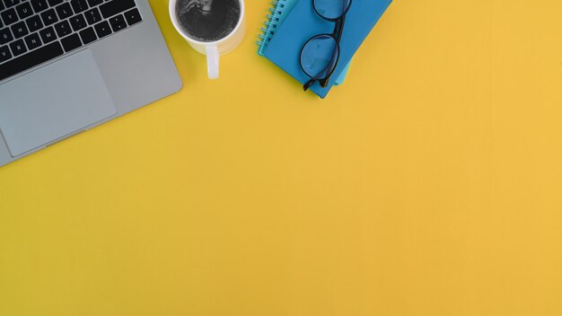 Bureaulaptop, koffie, notitieboekje en glazen op geel bureau met exemplaarruimte.