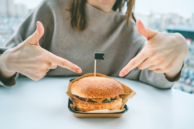 A burger in craft paper on a table with a flag and a girl points her fingers at it.