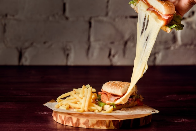 Burger in a cut with cutlet, melted cheese, bacon, lettuce, tomatoes and potatoes on parchment on a wooden platter on a wooden table.