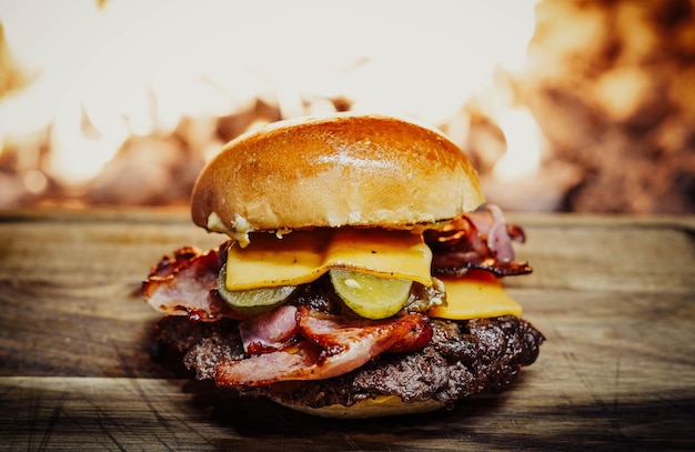 Foto un hamburger con formaggio, sottaceti e pancetta su un tavolo di legno accanto a un fuoco.