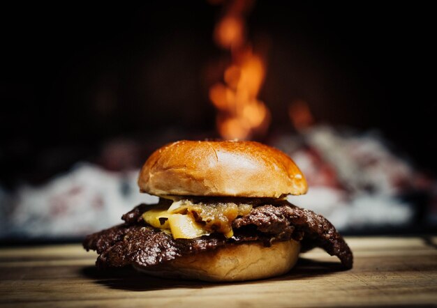 A burger with a grilled cheese on it sits on a wooden table with a fire in the background.