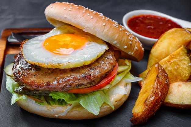 Burger with meat and eggs, on a board with sauce and potatoes