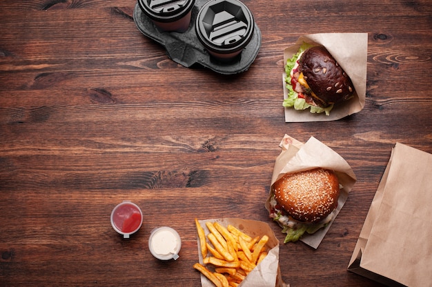 Burgers, french fries with sauces and coffee to go top view on wood. Horizontal shot.