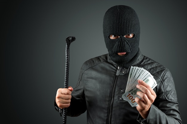 Burglar in a balaclava holds dollars in his hands on a dark background