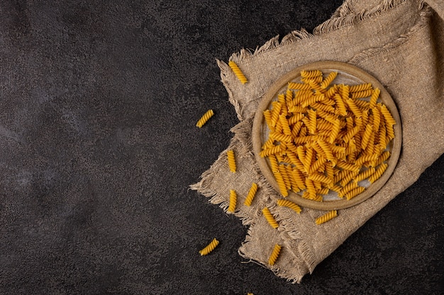 Burlap napkin on a dark structural background top view On it is a scattering of pasta in a ceramic plate Copyspace