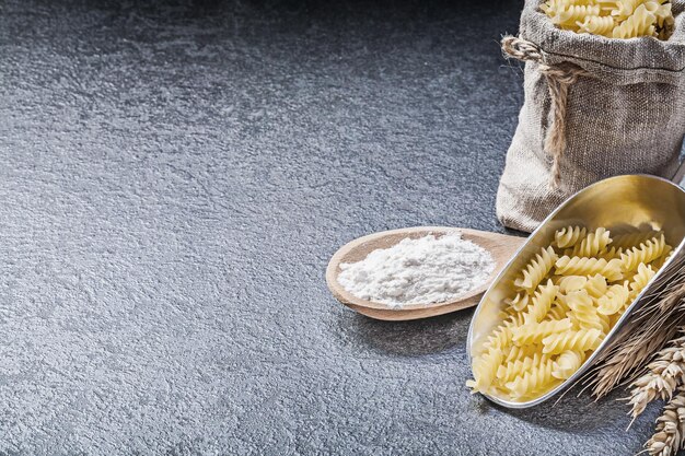 Burlap sack uncooked pasta kitchen scoop wooden spoon flour wheat rye ears on black background.