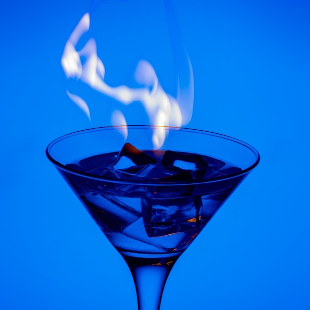 Burning alcoholic drink with ice cubes on a blue gradient\
background vertical frame burning cocktail on table in a bar blue\
alcoholic beverage and fire