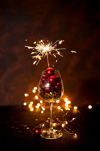 Burning bengal firework in wine glass with red Christmas balls on background of golden bokeh lights. Creative aesthetic moment. New Year atmosphere and mood at night. Selective focus