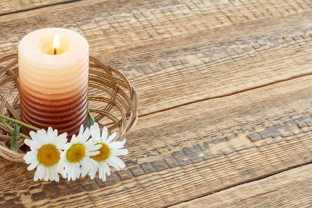Burning candle and bouquet of chamomile flowers in wicker basket on wooden boards. Top view. Holiday concept.