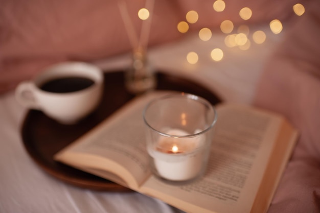 Burning candle staying on open paper book and cup of tea with\
aroma sticks at background over glowing lights. cozy atmosphere\
concept. good morning. home perfume. freshness.