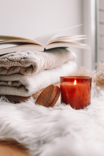 Burning candle and sweaters with a book on the windowsill. Autumn mood.