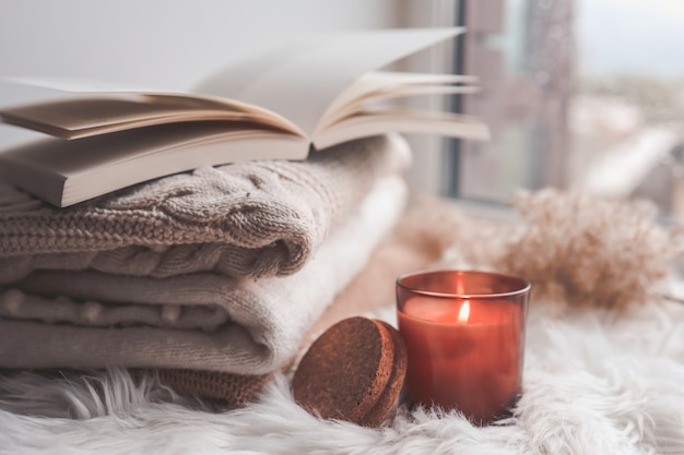 Burning candle and sweaters with a book on the windowsill. Autumn mood.