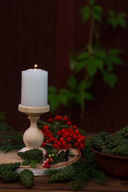 Горящая свеча на деревянном фоне. Деревянный стол украшен красной рябиной, зелеными сосновыми шишками и осенней листвой.