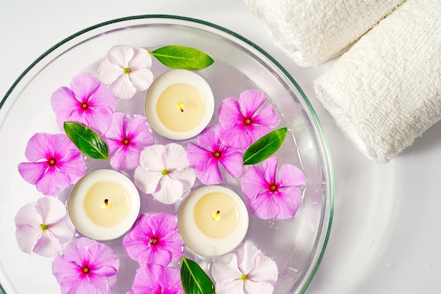 Burning floating candles with pink flowers in a bowl of water and two white towels spa relaxation bo...