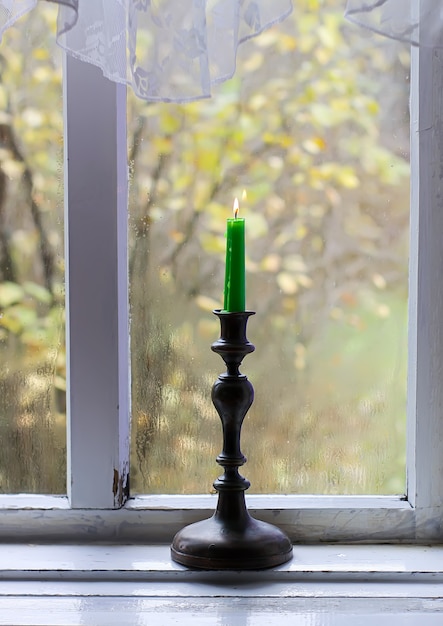 Burning wax candle in brass candlestick on the wooden window sill of in country house interior.