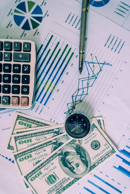 Business accessories (notebook, magnifier, calculator, planchette, tablet, fountain pen, notebook, glasses) and graphics, tables, charts on a wooden office desk. Soft focus