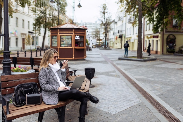 Donna caucasica adulta di affari che si siede con il computer portatile e che parla sul vivavoce nella piazza della città