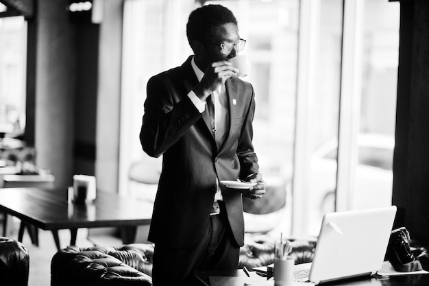 Foto l'uomo afroamericano d'affari indossa l'abito nero e gli occhiali in ufficio con il computer portatile che beve il caffè del mattino prima della giornata di lavoro