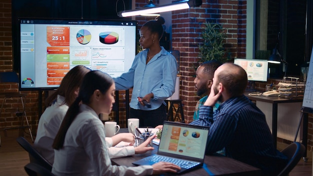 Business analytics meeting, african american project manager\
showing corporate presentation, team discussing statistics,\
planning sales strategy. coworkers watching diagrams at digital\
board