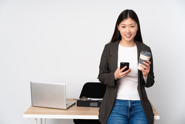 Business asian woman in her workplace isolated on white wall holding coffee to take away and a mobile