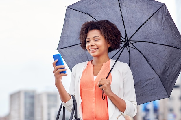 ビジネス、悪天候、人々、コンセプト - 街の通りでスマートフォンで傘のテキスト メッセージを送信する幸せなアフリカ系アメリカ人実業家