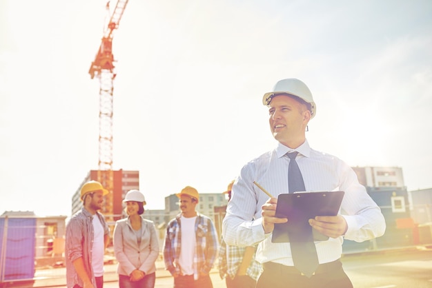 business, building, teamwork en people concept - groep lachende bouwers en architect met klembord in veiligheidshelmen op bouwplaats