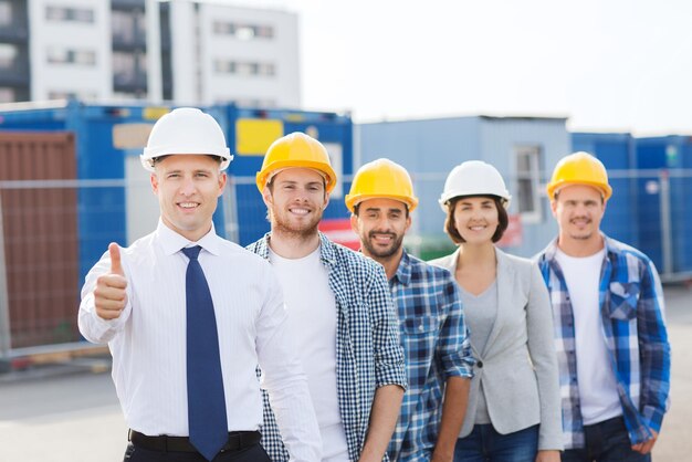 business, building, teamwork, gesture and people concept - group of smiling builders in hardhats showing thumbs up outdoors