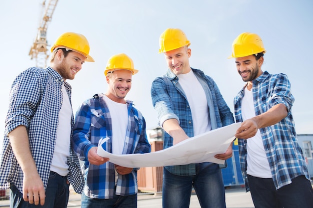 business, building, teamwork and people concept - group of smiling builders in hardhats with blueprint outdoors