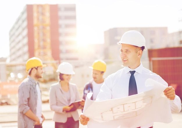 business, building, teamwork and people concept - group of smiling builders in hardhats with clipboard and blueprint outdoors