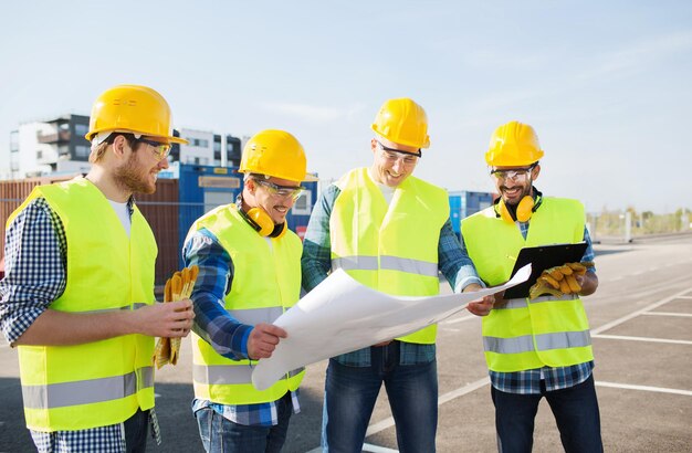 business, building, teamwork and people concept - group of smiling builders in hardhats with clipboard and blueprint outdoors