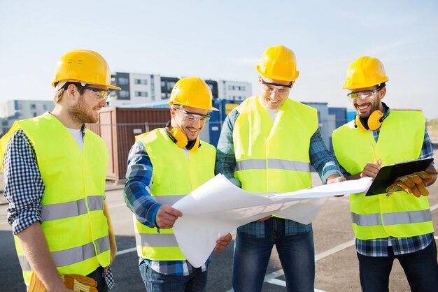 business, building, teamwork and people concept - group of smiling builders in hardhats with clipboard and blueprint outdoors