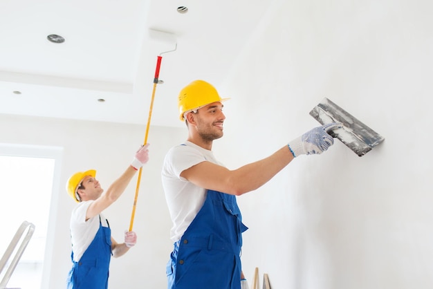 business, building, teamwork and people concept - group of smiling builders in hardhats with plastering tools indoors