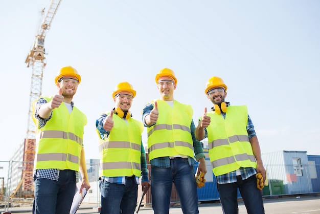 Concetto di affari, costruzione, lavoro di squadra, tecnologia e persone - gruppo di costruttori sorridenti in elmetti protettivi con computer tablet pc e appunti che mostrano i pollici in su gesto all'aperto