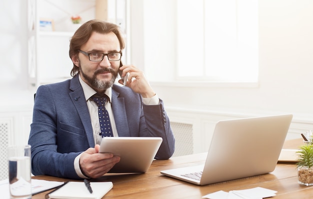 Business call. Successful serious businessman talking on mobile phone and working on laptop in modern white office, copy space