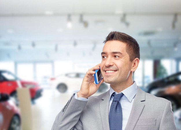 Business, car sale, people and technology concept -happy\
businessman calling on smartphone over auto show or salon\
background