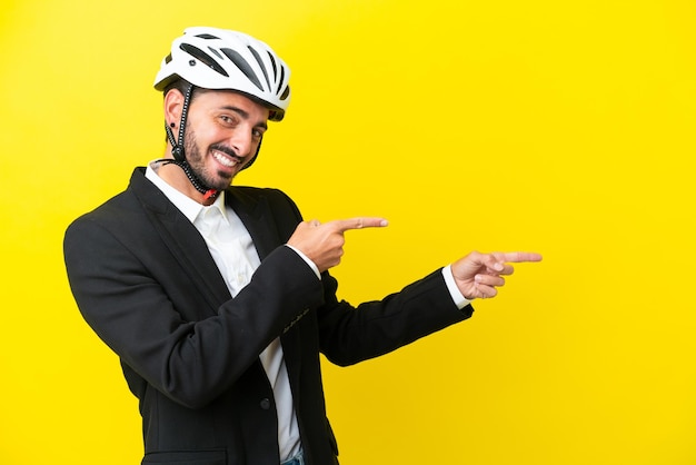 横に指を指し、製品を提示する黄色の背景に分離された自転車のヘルメットを持つビジネス白人男性