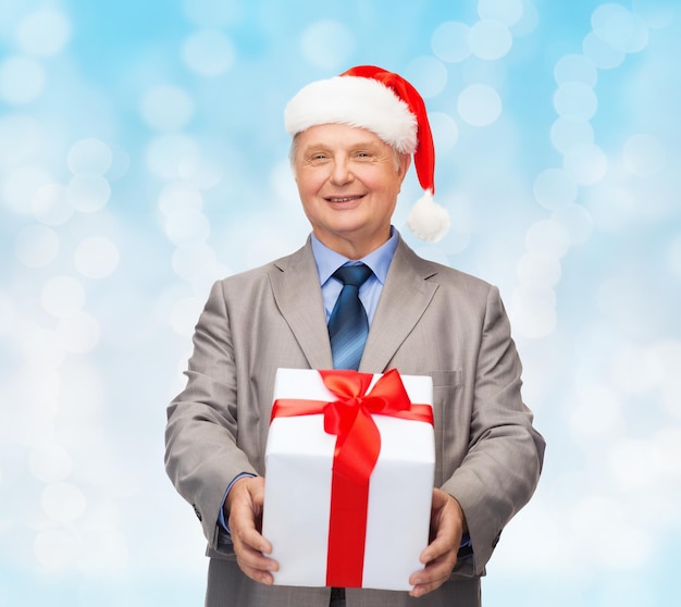 business, christmas, presents and people concept - smiling senior man in suit and santa helper hat with gift over blue lights background