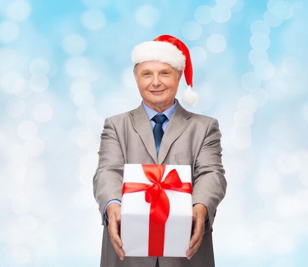 business, christmas, presents and people concept - smiling senior man in suit and santa helper hat with gift over blue lights background