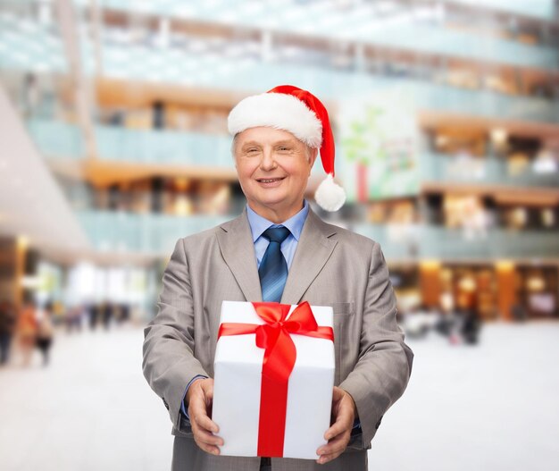 Concetto di affari, natale, regali e persone - uomo anziano sorridente in tuta e cappello da assistente di babbo natale con regalo su sfondo centro commerciale