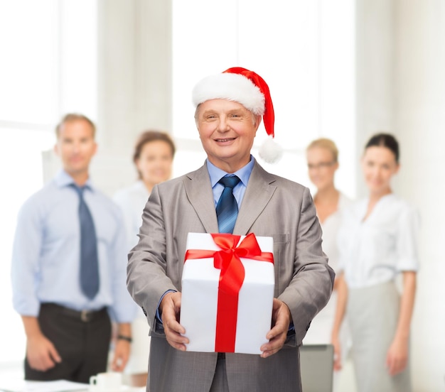 Photo business, christmas, xmas, happiness concept - smiling old man in suit and santa helper hat with gift