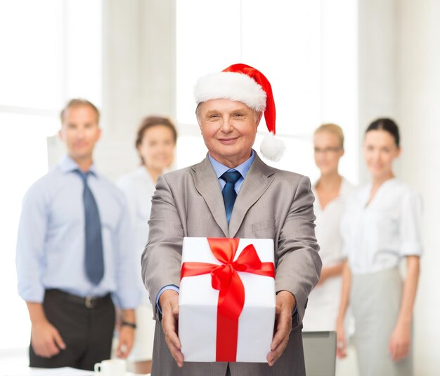 Photo business, christmas, xmas, happiness concept - smiling old man in suit and santa helper hat with gift